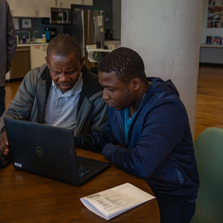 s wearing a gray sweater over a light blue shirt, while the younger individual on the right is wearing a blue hoodie. Both are focused on the laptop screens. In the background, a brightly lit room features a kitchen area and a wall with photos. A printed document is on the table in front of them.