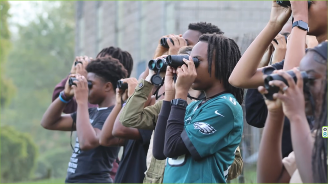 A picture of students at Lankenau Environmental Science Magnet High School look through binoculars during a learning activity outside the school building.